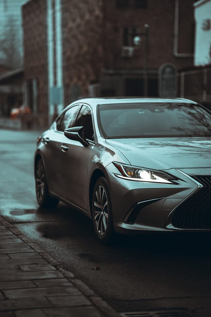 Silver sedan with glowing headlights parked on a wet urban street with blurred brick buildings in the background.