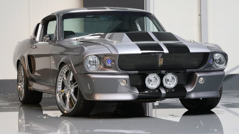 Gray Shelby GT500 muscle car with black racing stripes and chrome wheels on a reflective studio floor.