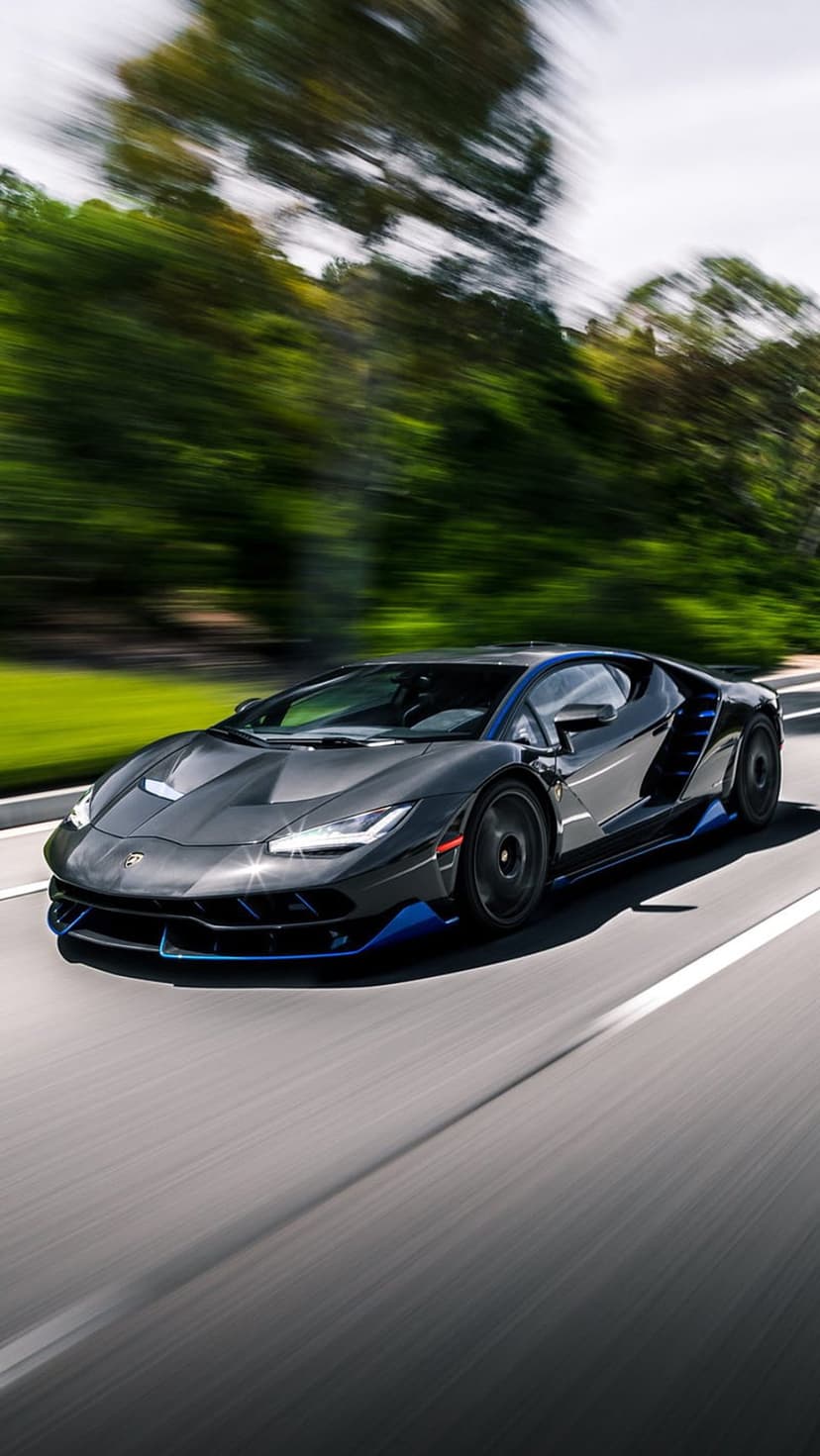 Lamborghini Centenario with blue accents speeds on a highway, flanked by motion-blurred green trees and bushes.