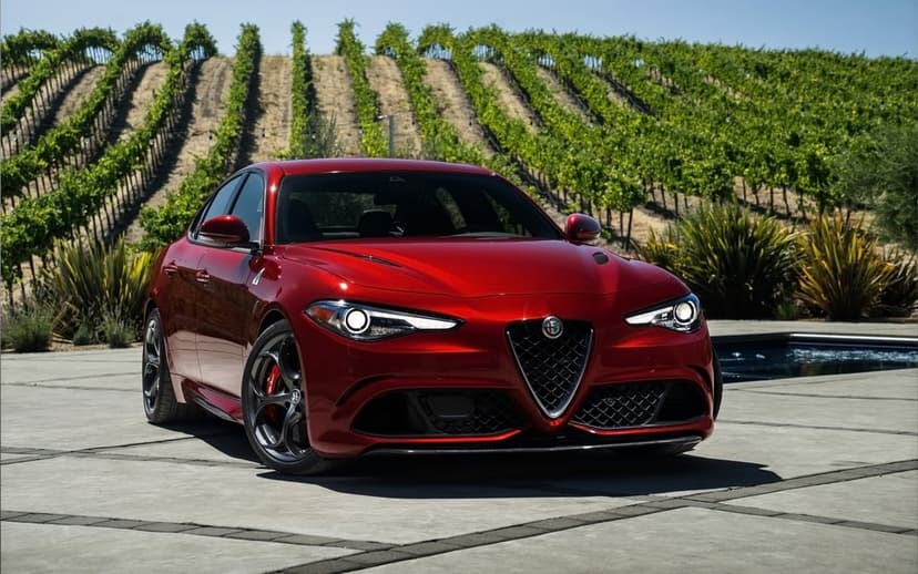 Red Alfa Romeo sports sedan parked on concrete with a lush green vineyard and sunny hills in the background.