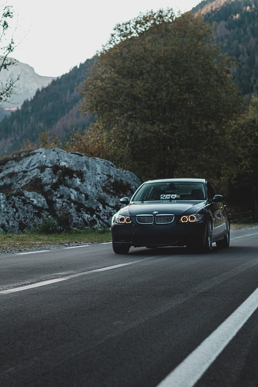 Dark BMW sedan with headlights on driving through a rocky autumn mountain pass surrounded by dense pine trees.