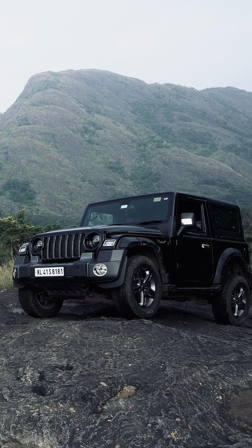 Black Mahindra Thar SUV parked on a jagged rock formation overlooking foggy green mountains and hazy skies.