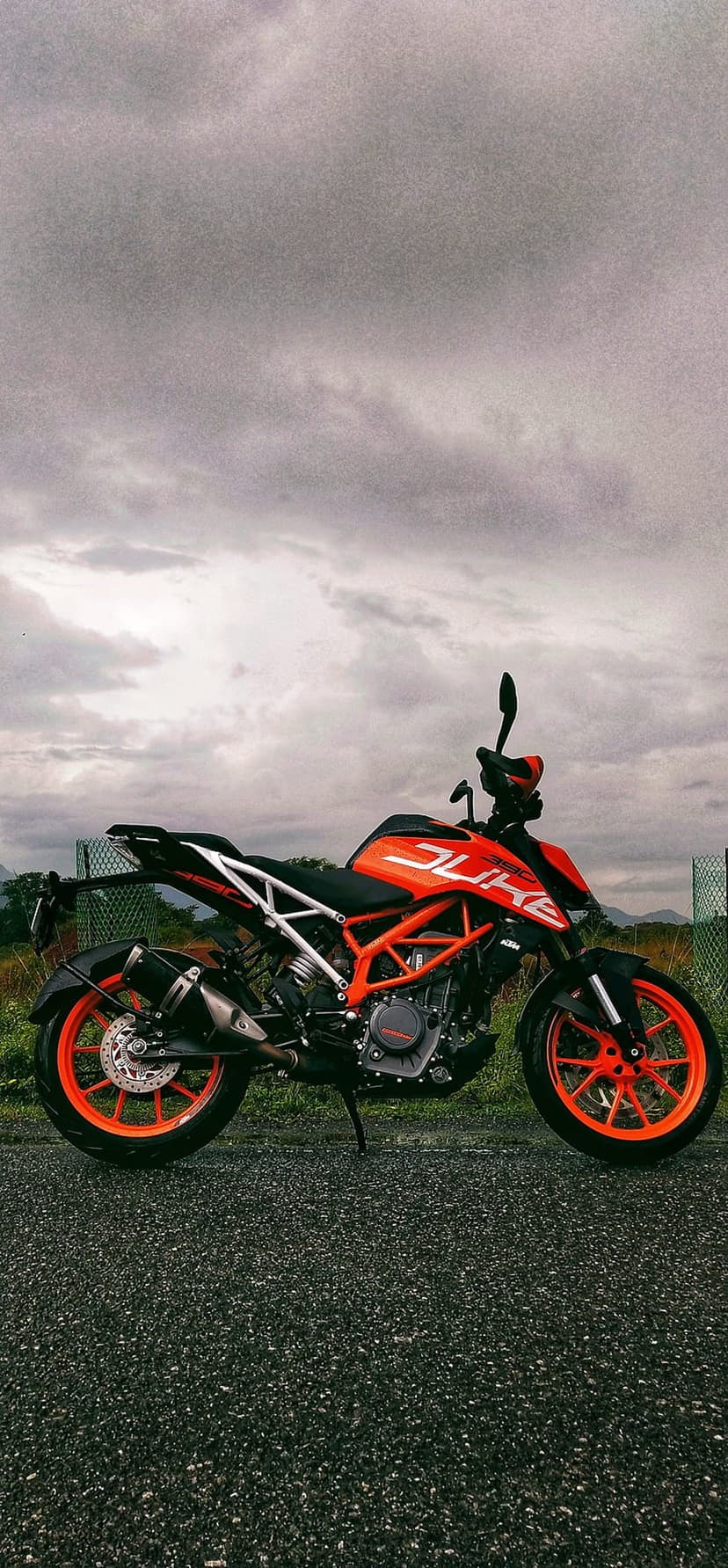 KTM Duke motorcycle with orange frame parked on dark asphalt under a moody sky with distant mountains and fields.