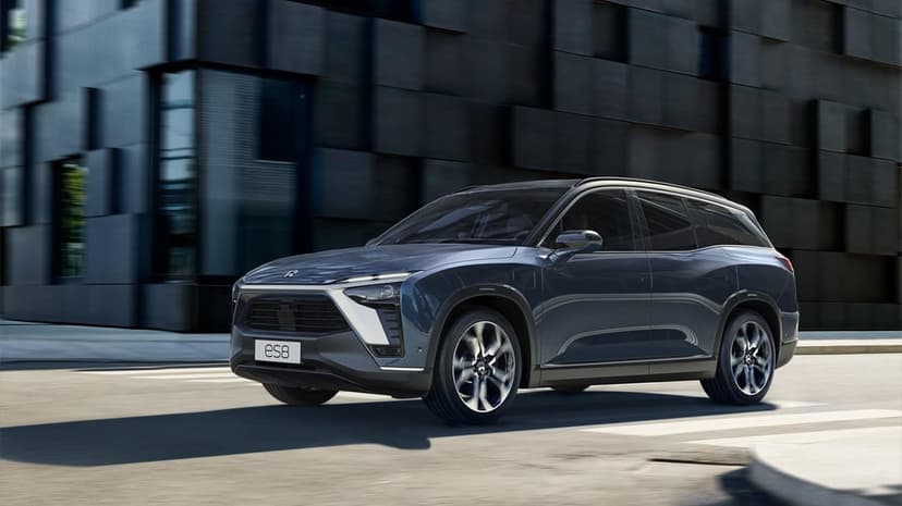 Dark blue NIO ES8 electric SUV driving on a city street beside a modern textured charcoal building facade.