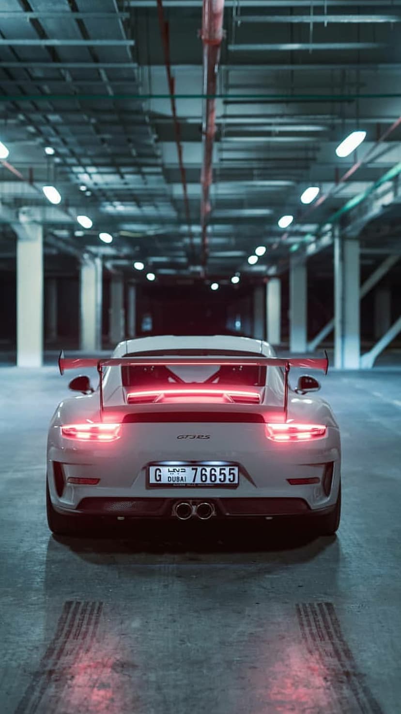 White Porsche GT3 RS rear view showing glowing red taillights and large spoiler in a dim Dubai parking structure.