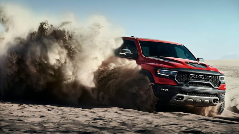 Red Ram TRX pickup truck driving through sand dunes creating a large dust cloud under a clear blue sky.