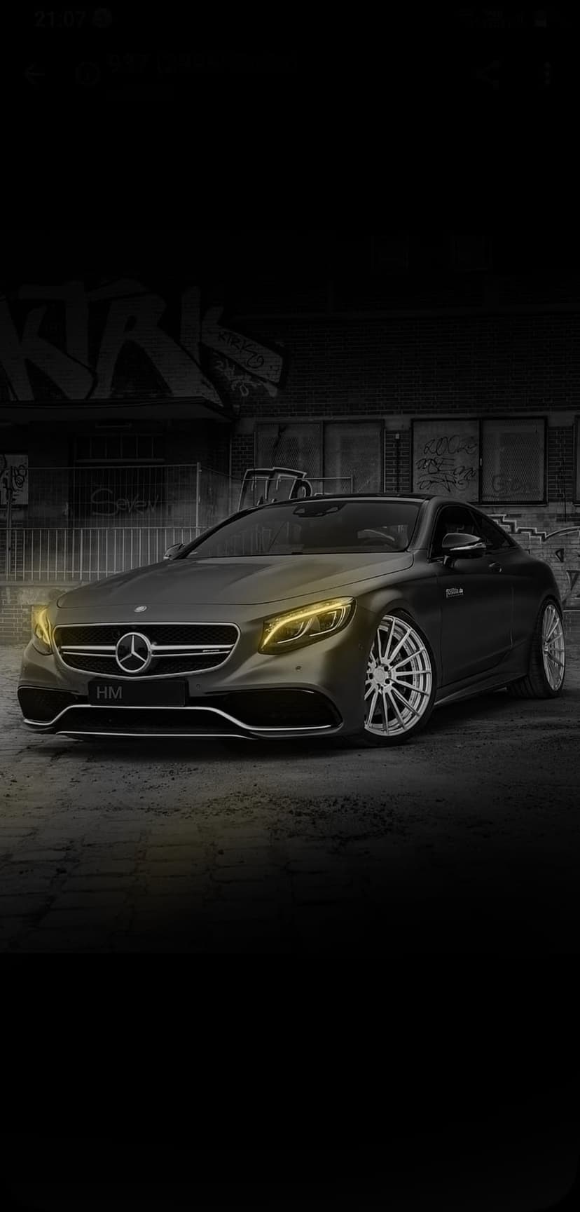 Dark Mercedes S-Class coupe with yellow headlights parked in front of a colorful urban graffiti mural on a street.