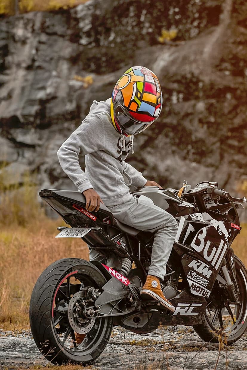 Motorcyclist in a colorful helmet sits on a black KTM bike against a rocky off-road background with dry grass.