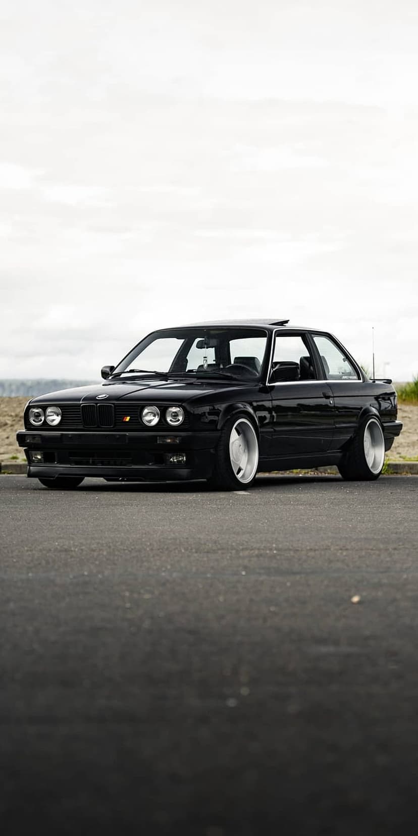 Black BMW E30 coupe with white deep-dish wheels parked on asphalt against a blurred ocean and blue sky horizon.