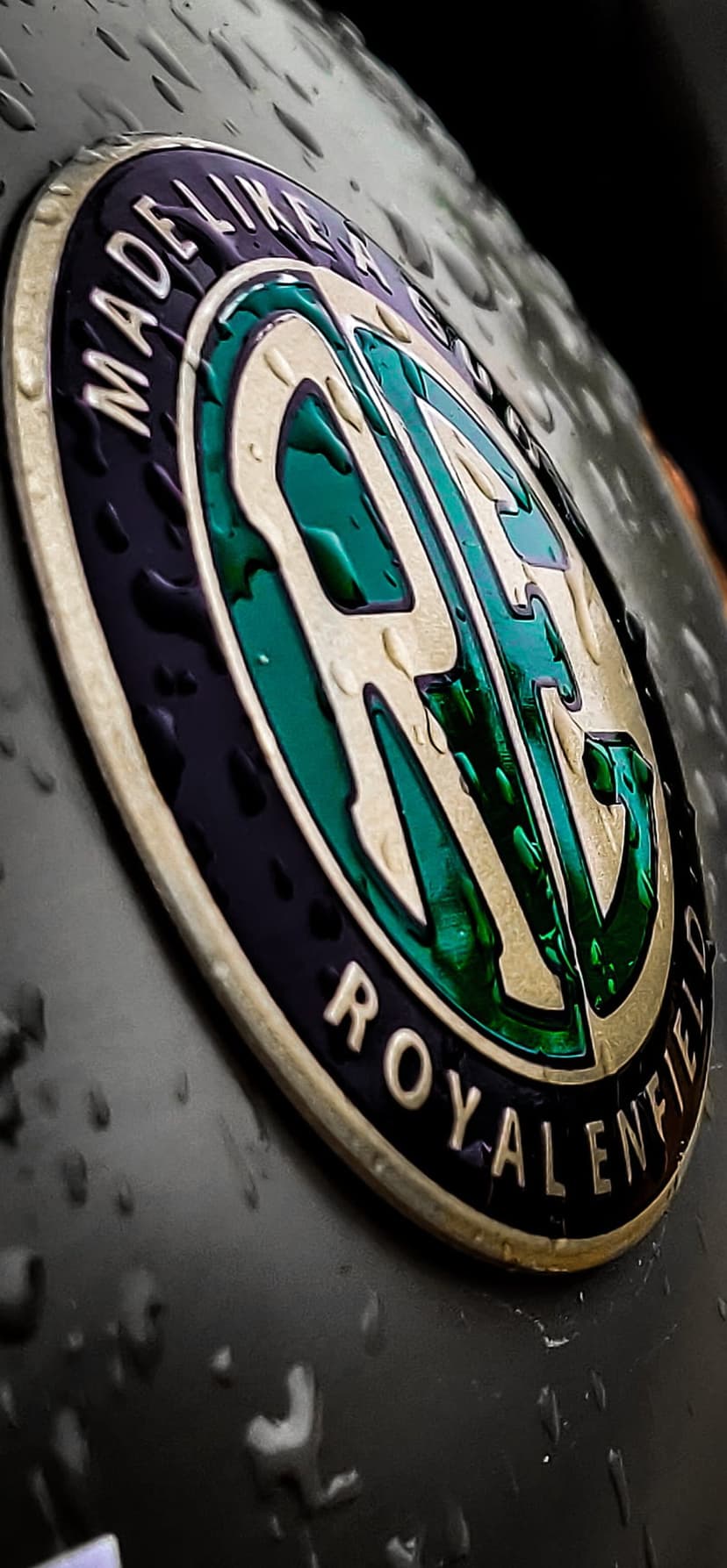 Royal Enfield chrome emblem with water droplets and Made Like A Gun text on a dark metallic motorcycle tank.
