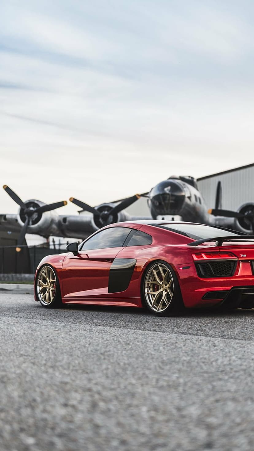 Red Audi R8 with gold rims parked on asphalt in front of a silver vintage propeller aircraft under a cloudy sky.