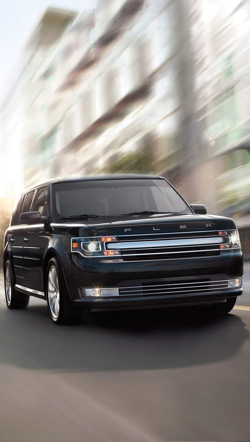 Black Ford Flex SUV with shining chrome grille moves through a blurred sunset city street with golden lighting.
