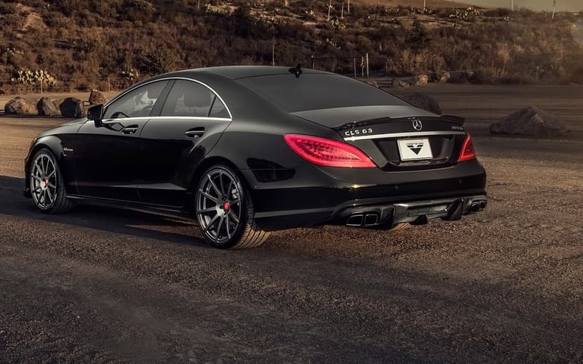 Rear view of a black Mercedes-Benz CLS 63 AMG parked on a dirt road during sunset with hills and cacti behind.