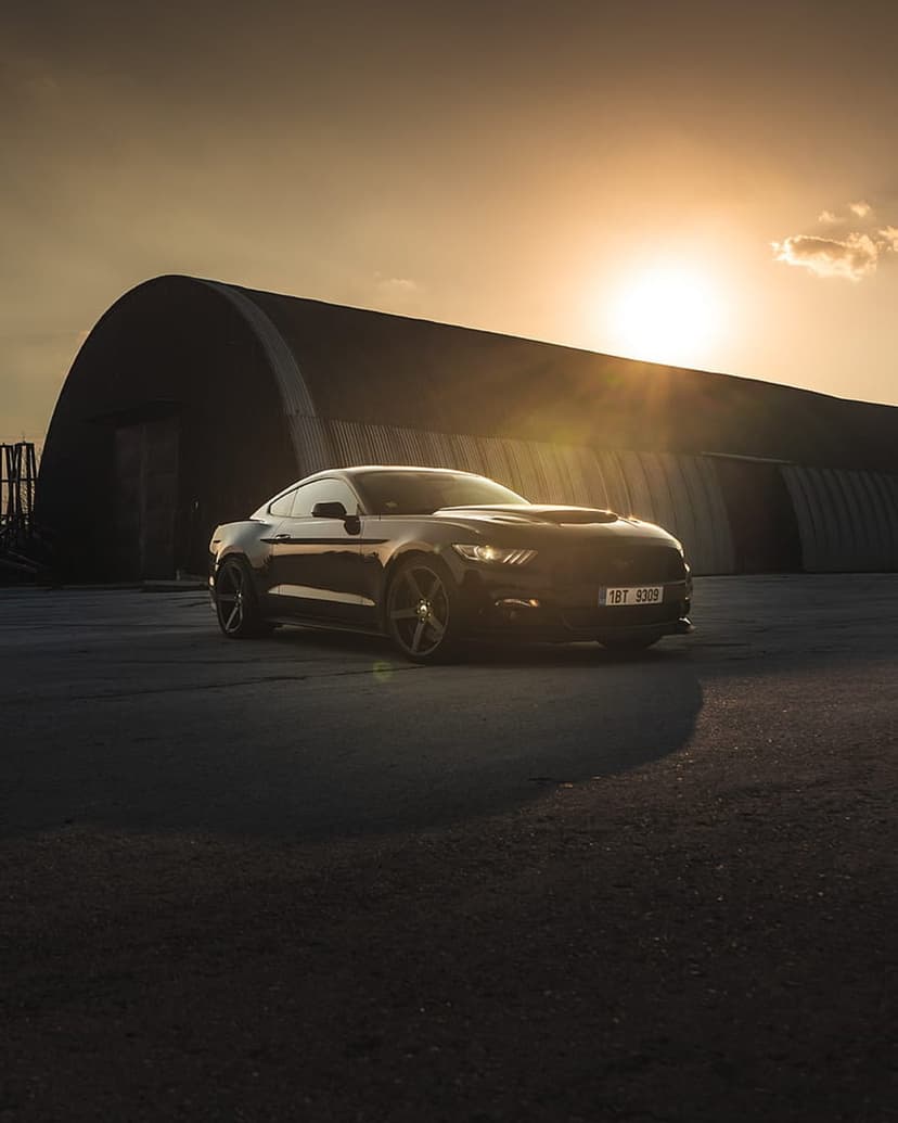 Black Ford Mustang parked on asphalt near a dark hangar building during a bright golden hour sunset with lens flare.