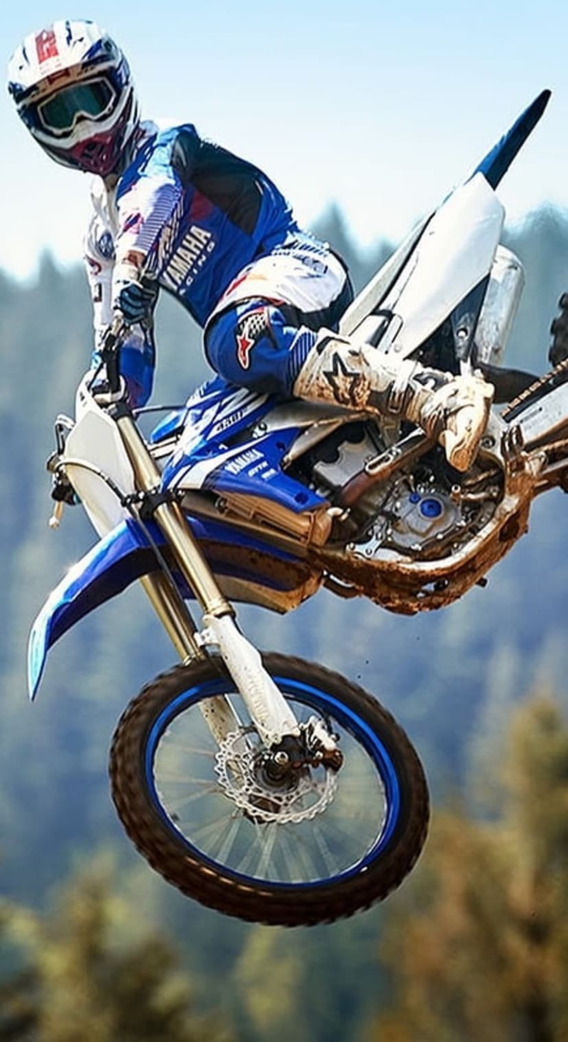 Motocross rider on a blue Yamaha dirt bike jumps mid-air against a blurred green forest background in full gear.