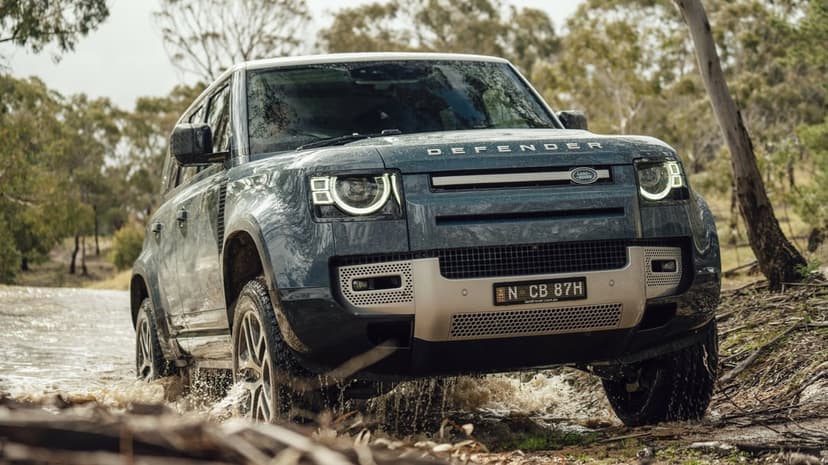 Blue Land Rover Defender driving through a shallow muddy stream with splashing water and forest background.
