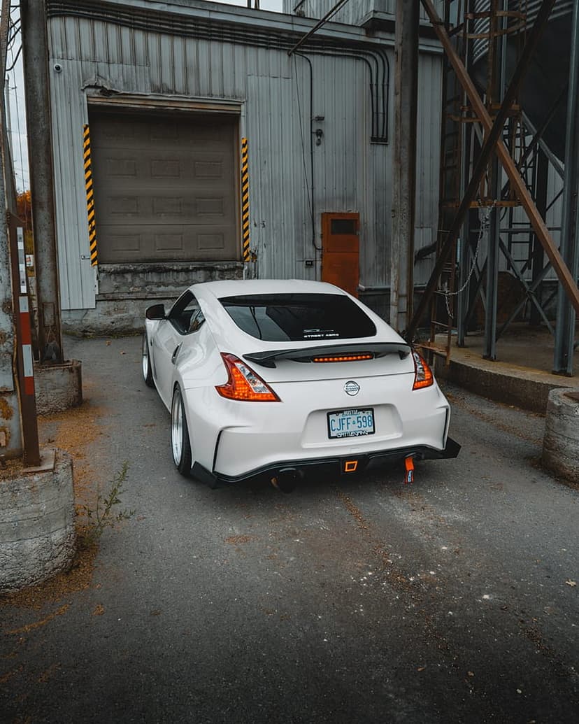 White Nissan 370Z with an aggressive body kit and orange tow hooks parked in an industrial warehouse bay.