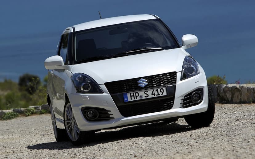 Front view of a white Suzuki Swift Sport on a gravel road with a deep blue sea and sky background in the distance.