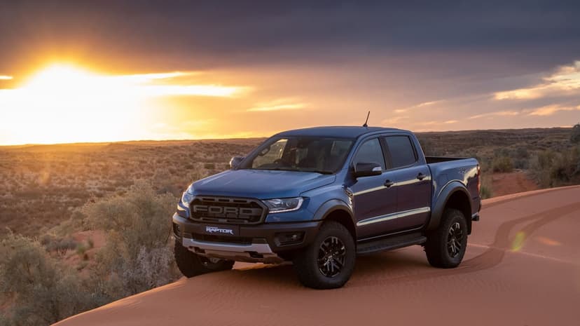 Blue Ford Ranger Raptor pickup truck on a desert sand dune during a vibrant golden hour sunset with orange skies.
