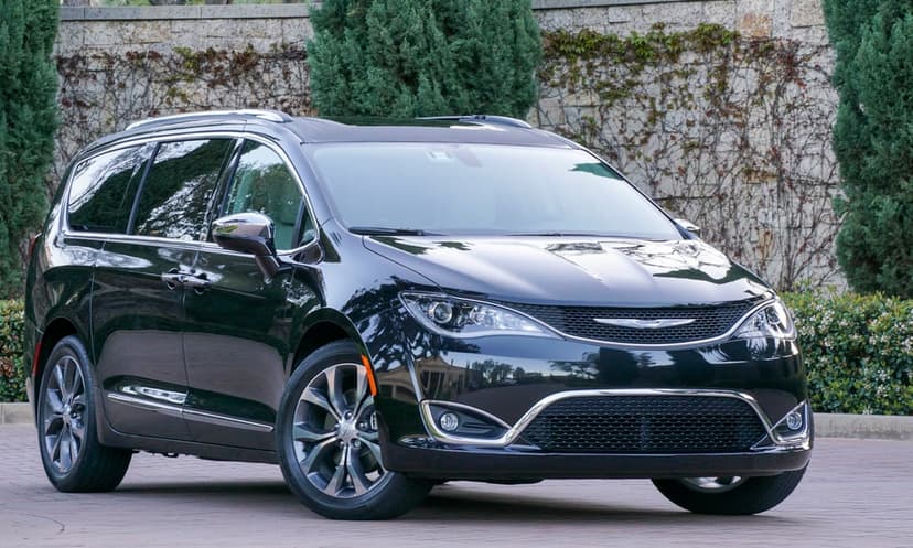 Black Chrysler Pacifica minivan parked on a paved driveway in front of a stone wall and green trees.