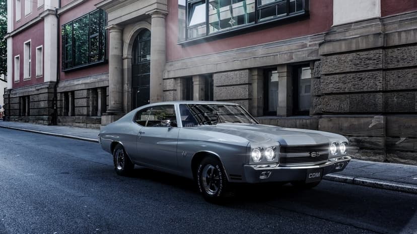 Silver 1970 Chevrolet Chevelle SS parked on a street in front of a stone building with glowing headlights.