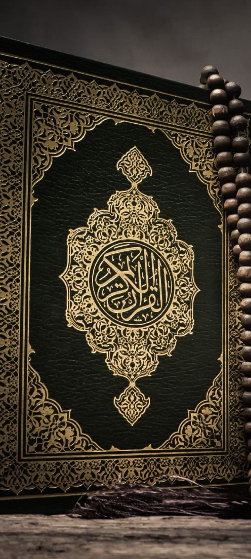 Intricate gold calligraphy on a green leather Quran next to a string of wooden prayer beads in low light.