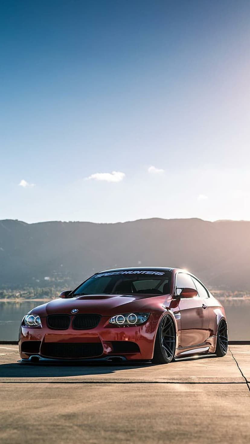 Red BMW M3 coupe with wide body fenders parked outdoors against a scenic mountain and lake horizon.