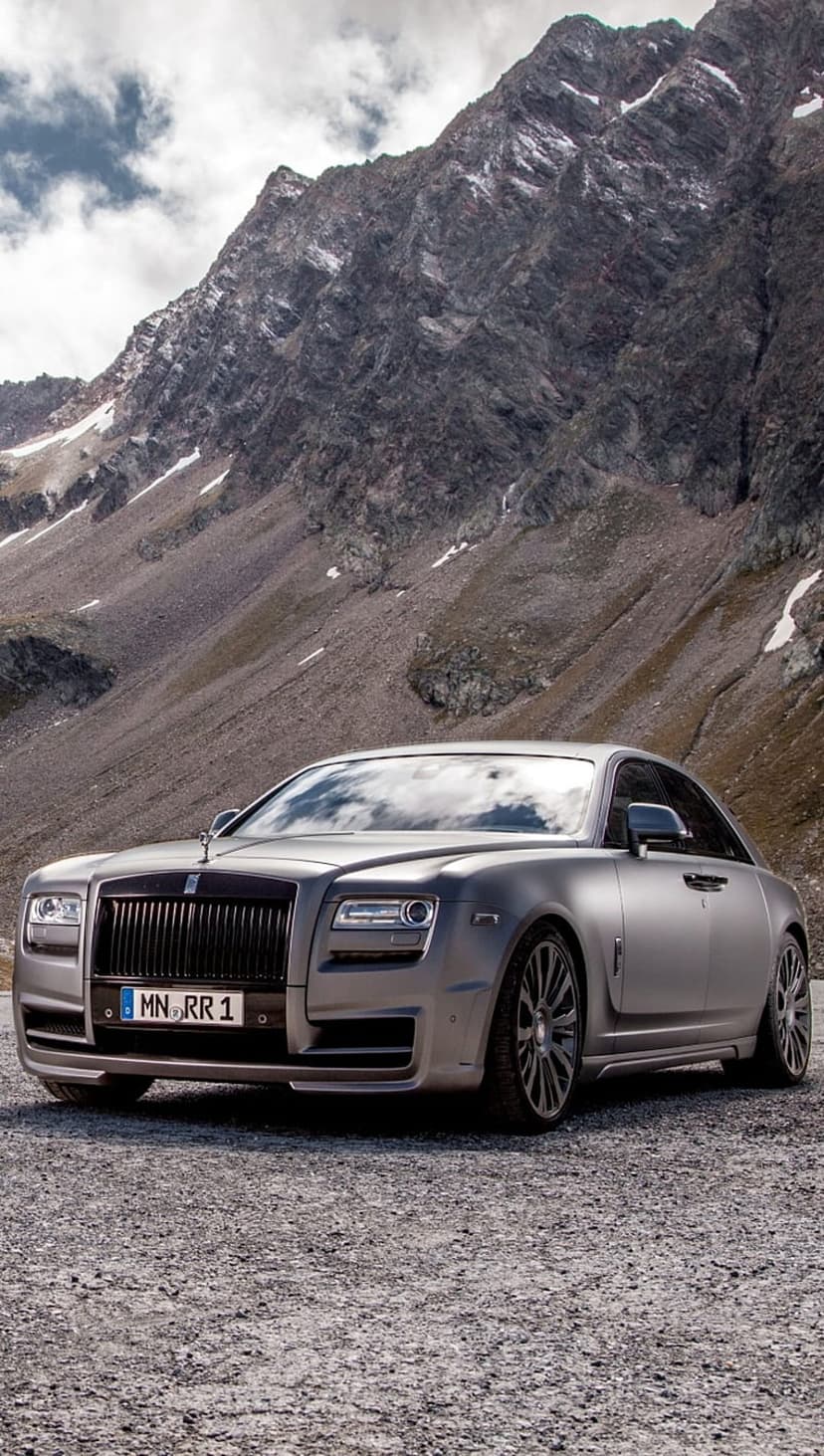 Rolls-Royce Ghost sedan in matte silver parked on a gravel mountain road under a cloudy sky during the day.