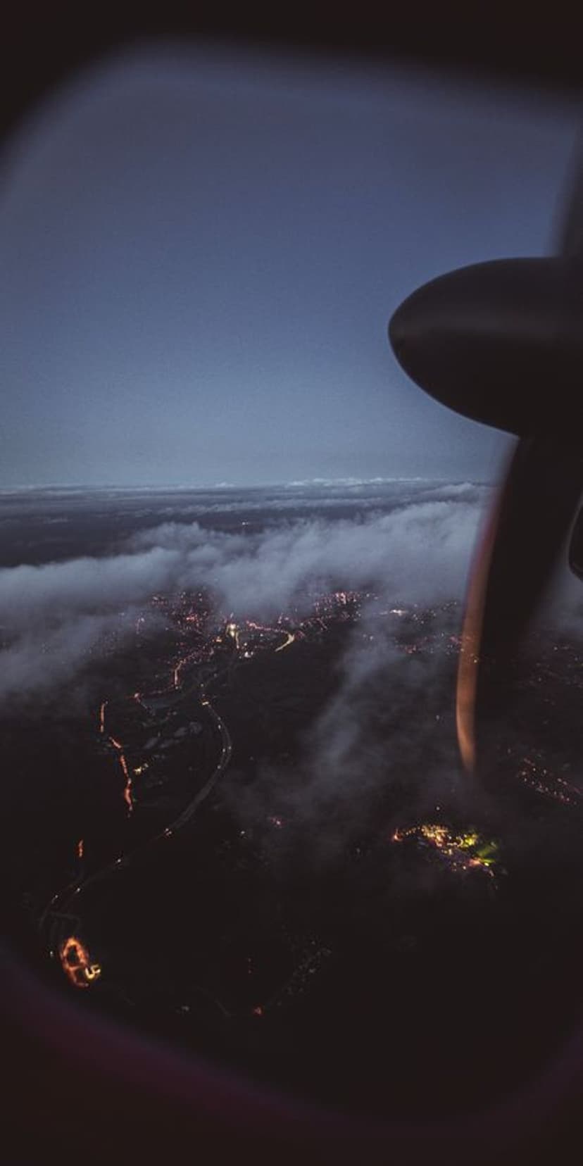 Glowing city lights shimmer through white clouds as seen from an airplane window with a blurred propeller.