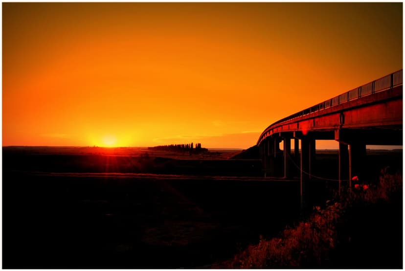 Curved bridge silhouette stretching across dark plains under a vibrant orange and red sunset sky at twilight.