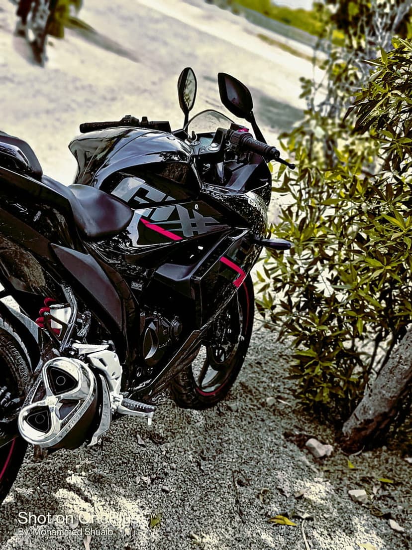 Black sportbike motorcycle with red highlights parked on gravel surrounded by dense green forest foliage and trees.