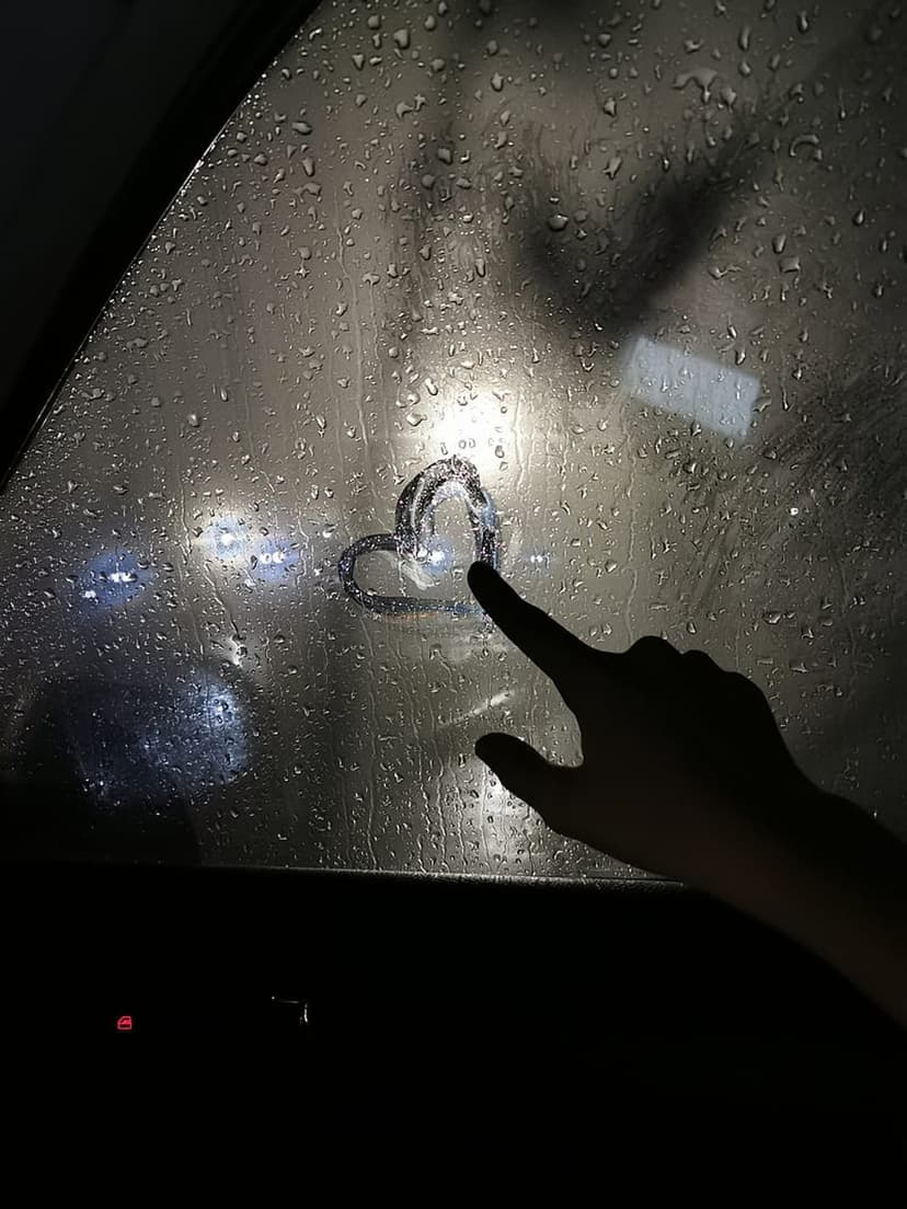Heart shape traced on a car window covered in raindrops with blurred golden streetlights in the background.
