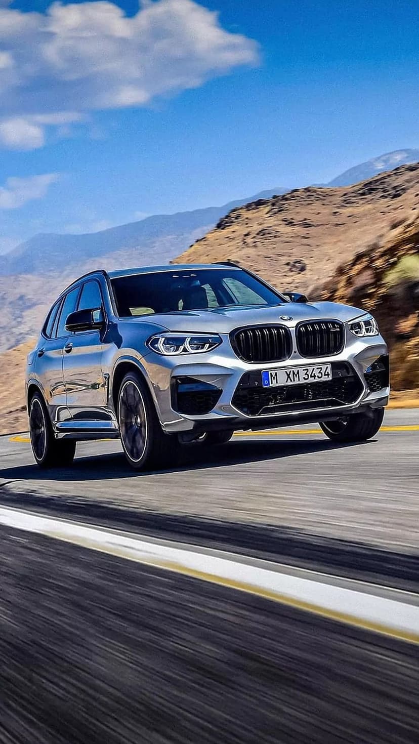 Silver BMW X3 M SUV accelerating on a winding mountain road with motion blur under a clear blue sky.