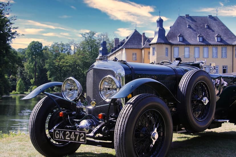 Dark green vintage Bentley with chrome accents parked on a lawn before a tall stone castle with many windows.