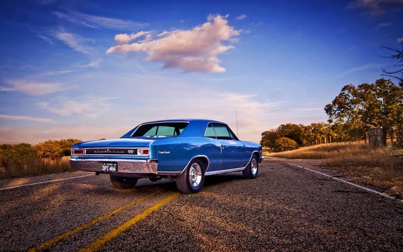 Rear view of a metallic blue 1966 Chevelle SS muscle car on a rural asphalt road under a sunset sky.