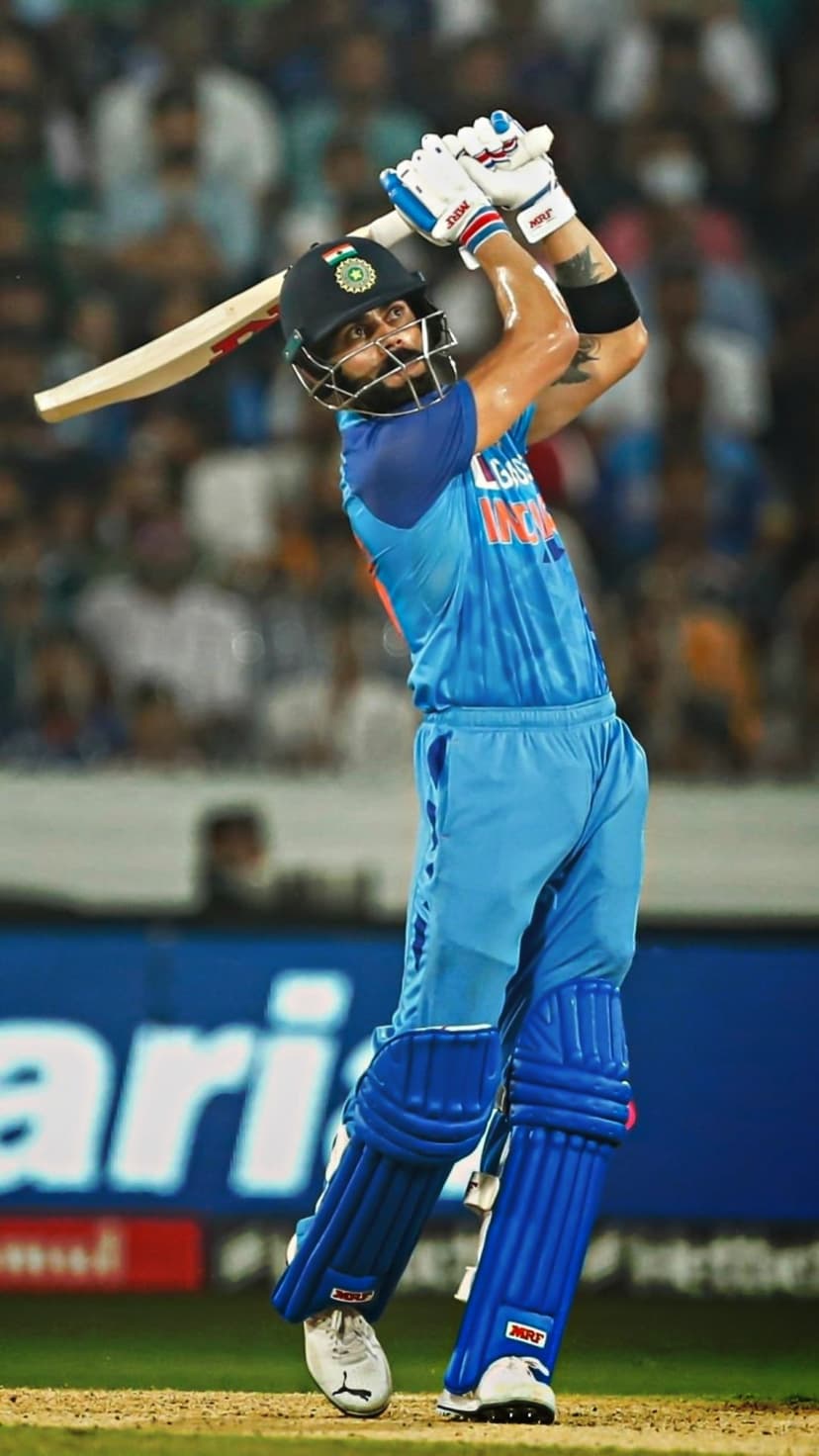 Virat Kohli swings his cricket bat in a blue team jersey while batting against a blurred stadium background.