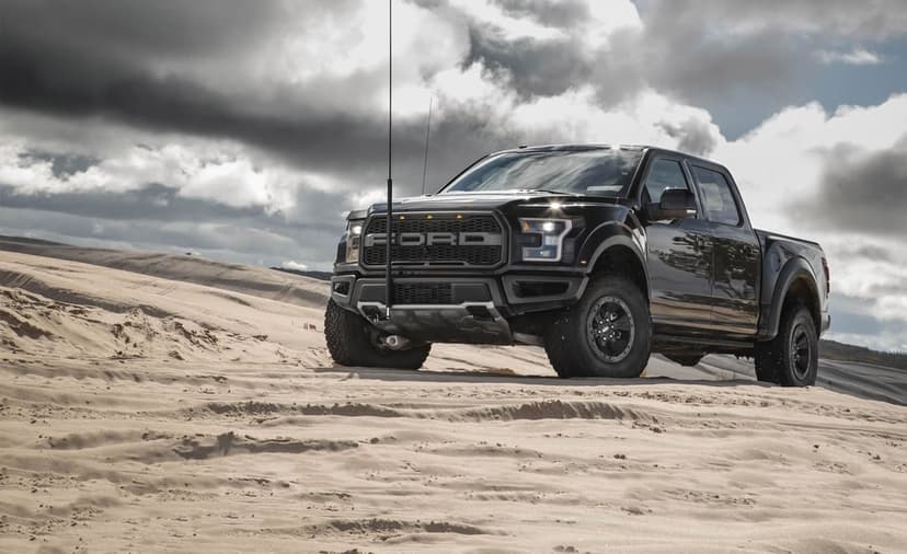 Black Ford Raptor truck on rippled sand dunes under dark, dramatic storm clouds in a landscape view.