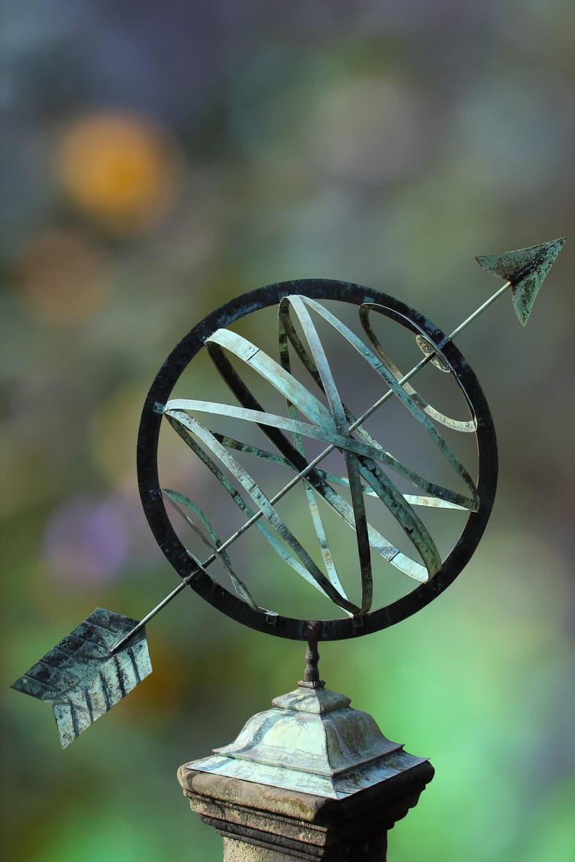 Bronze armillary sphere sundial with an arrow sits on a stone pedestal against a blurred garden backdrop.
