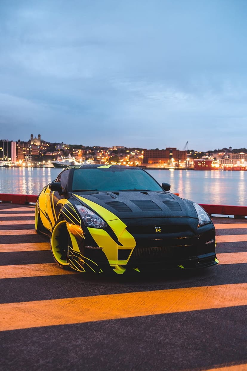 Yellow Nissan GT-R with black racing stripes parked on a pier with a glowing city skyline under a dusky sky.