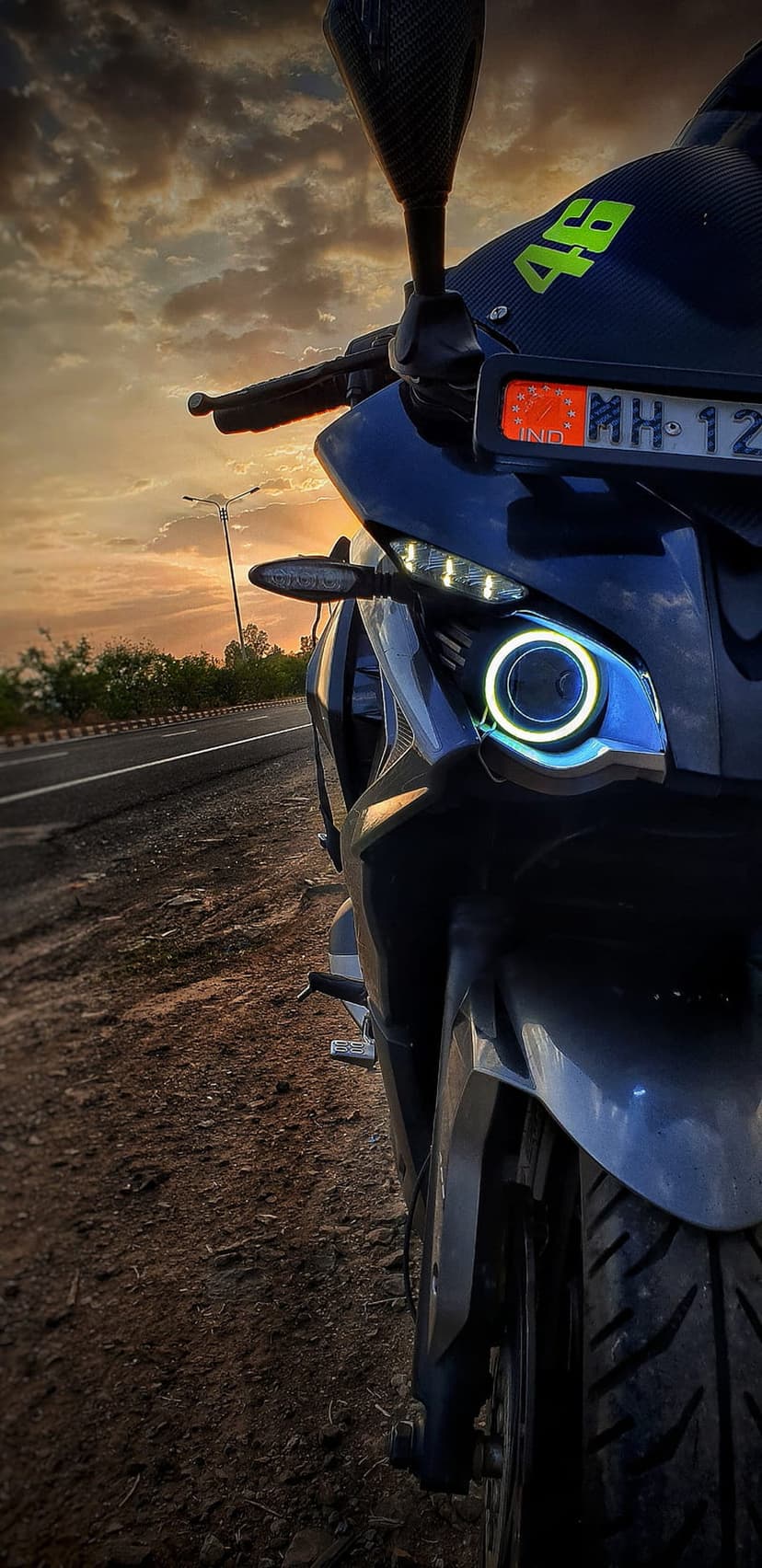 Motorcycle headlight and MH-12 license plate glow under an orange sunset sky with a background street lamp.