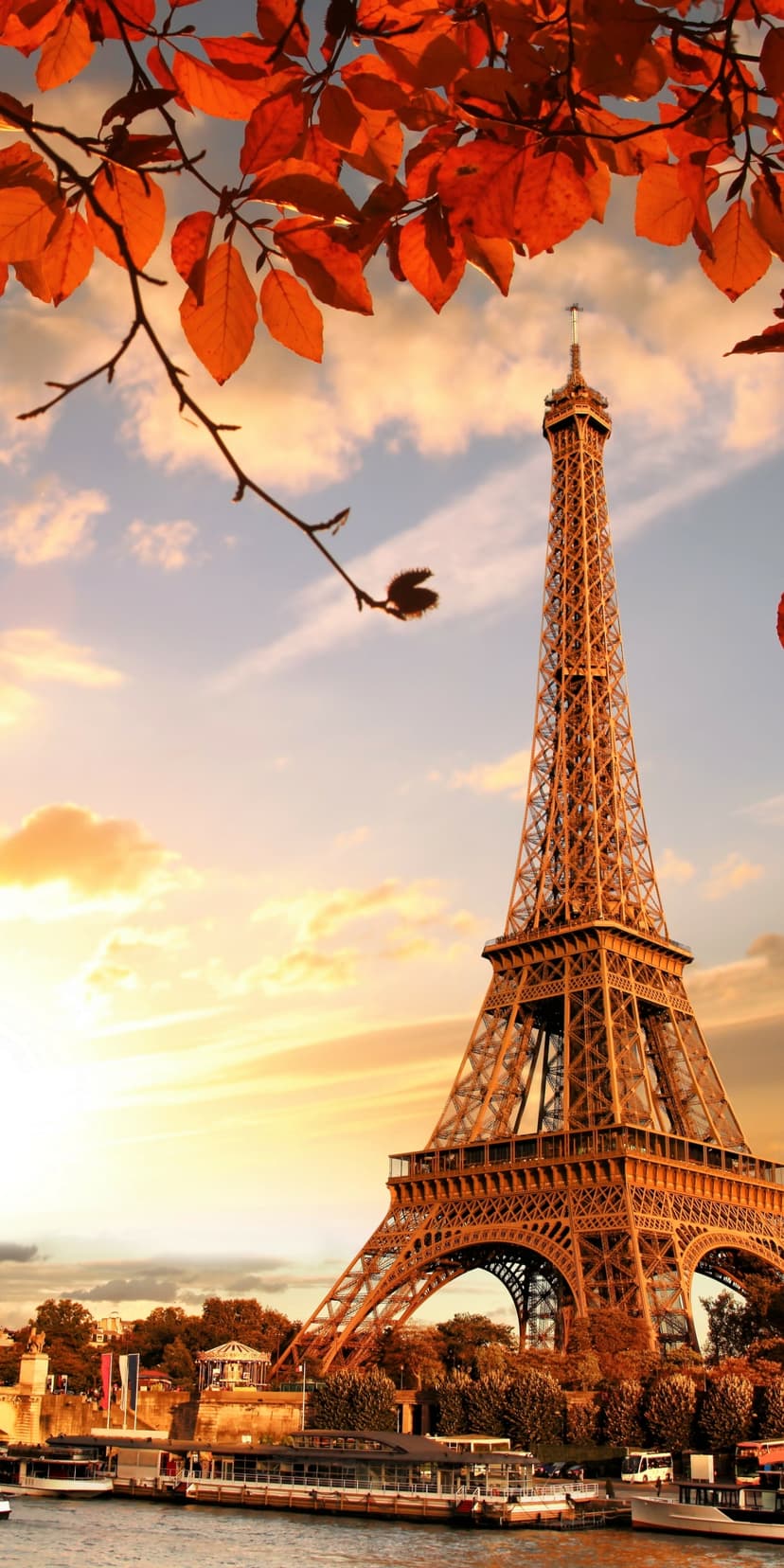 Eiffel Tower stands amidst orange autumn leaves and the Seine River under a glowing golden sunset sky.