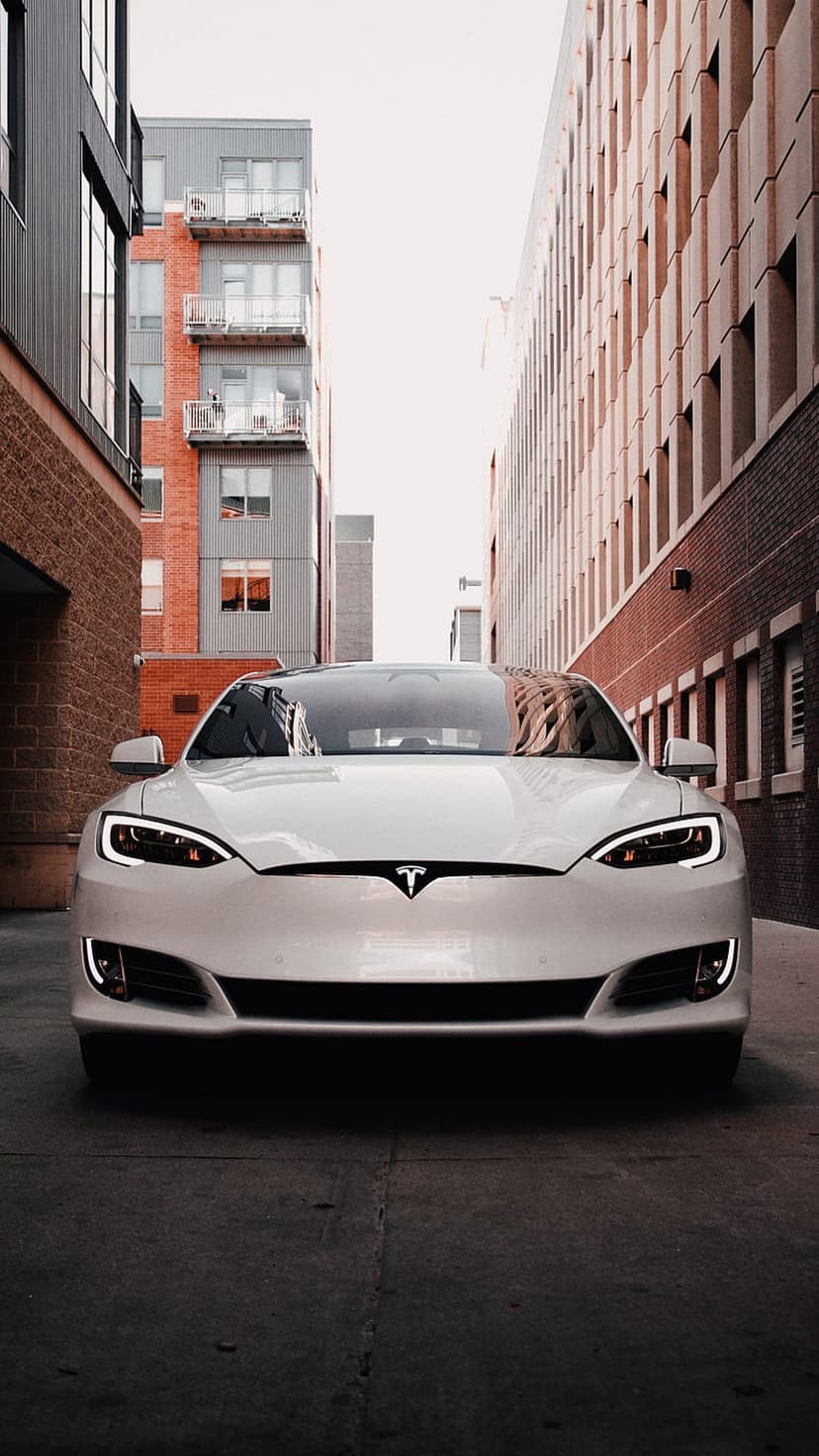 White Tesla sedan with glowing headlights parked in a narrow alleyway between industrial red brick buildings.