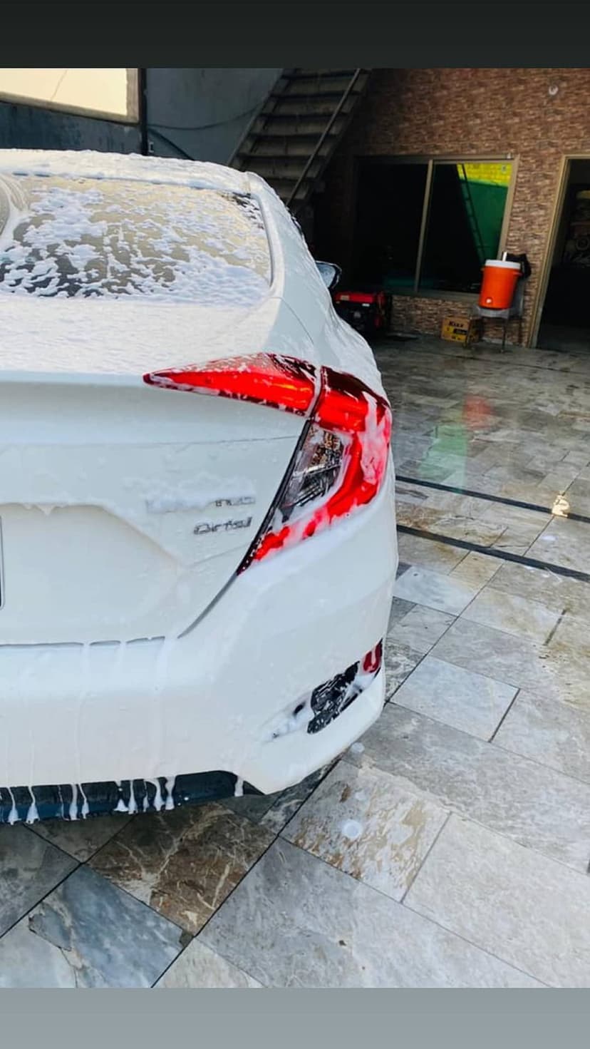 White car rear bumper and red taillights coated in thick white soap foam during an outdoor car wash session.