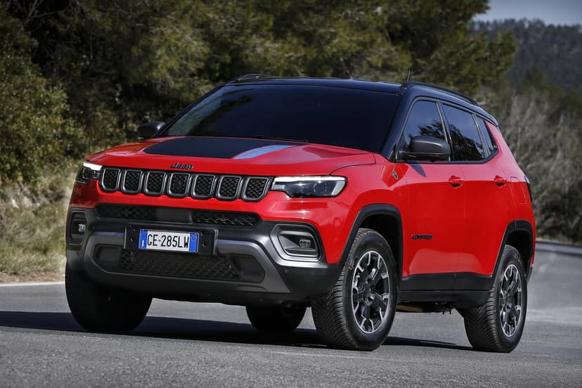Red Jeep Compass Trailhawk SUV with a black hood decal on a paved road surrounded by blurred green trees.