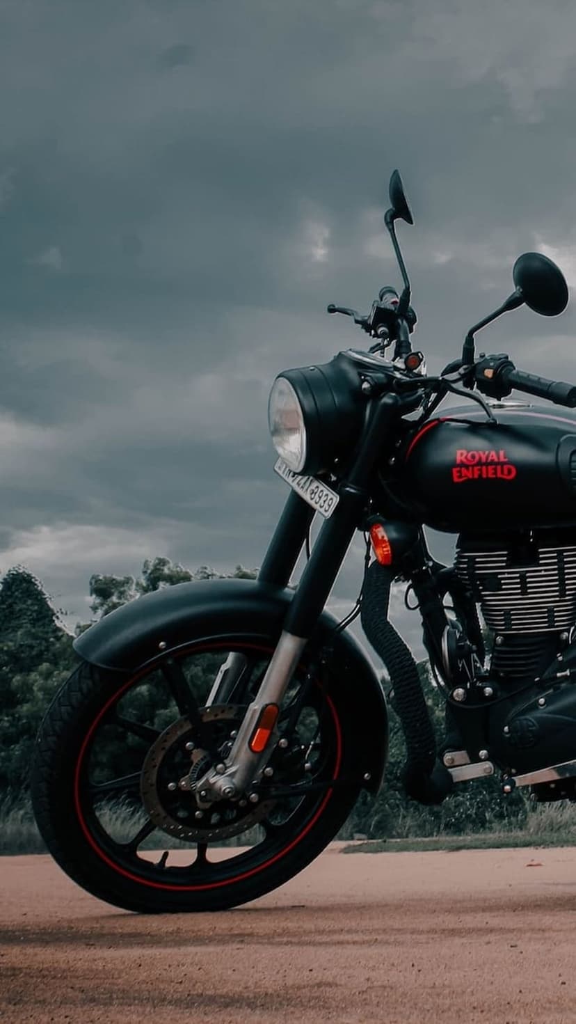 Black Royal Enfield motorcycle with a red logo parked on a dirt trail beneath a dark, heavy overcast sky.