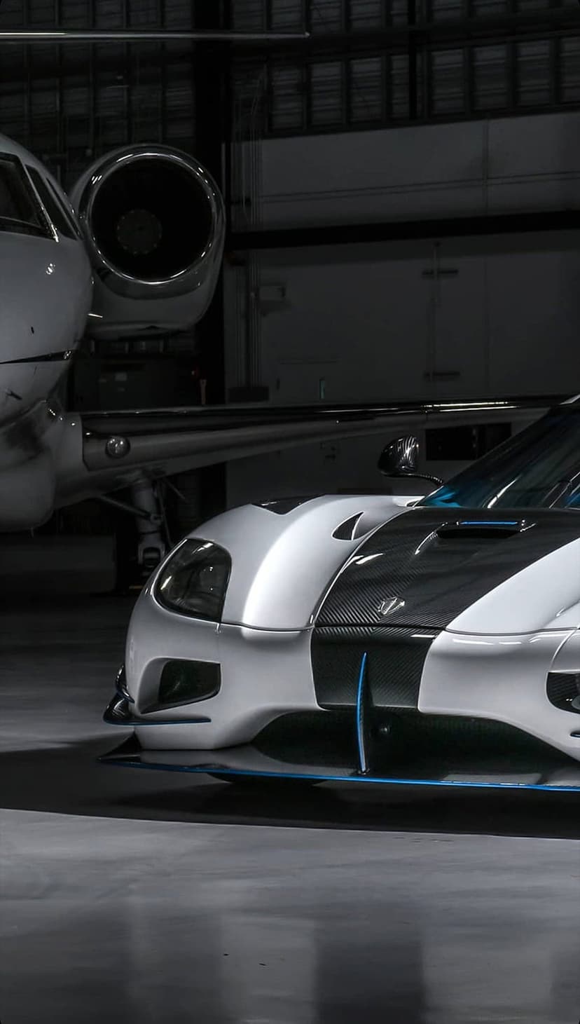 White sports car with carbon fiber accents parked next to a private jet in a dimly lit aviation hangar.