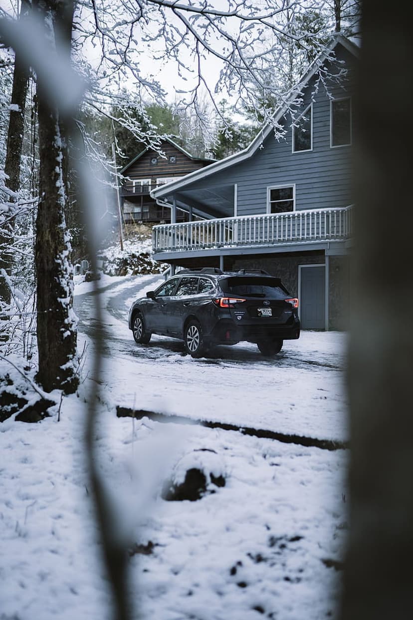 Dark SUV driving on a snowy road toward a modern gray house framed by frost-covered tree branches in a winter forest.