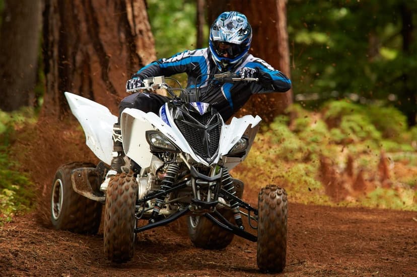 Person riding a white Yamaha ATV through a forest, kicking up dirt during an aggressive turn on a pine needle trail.