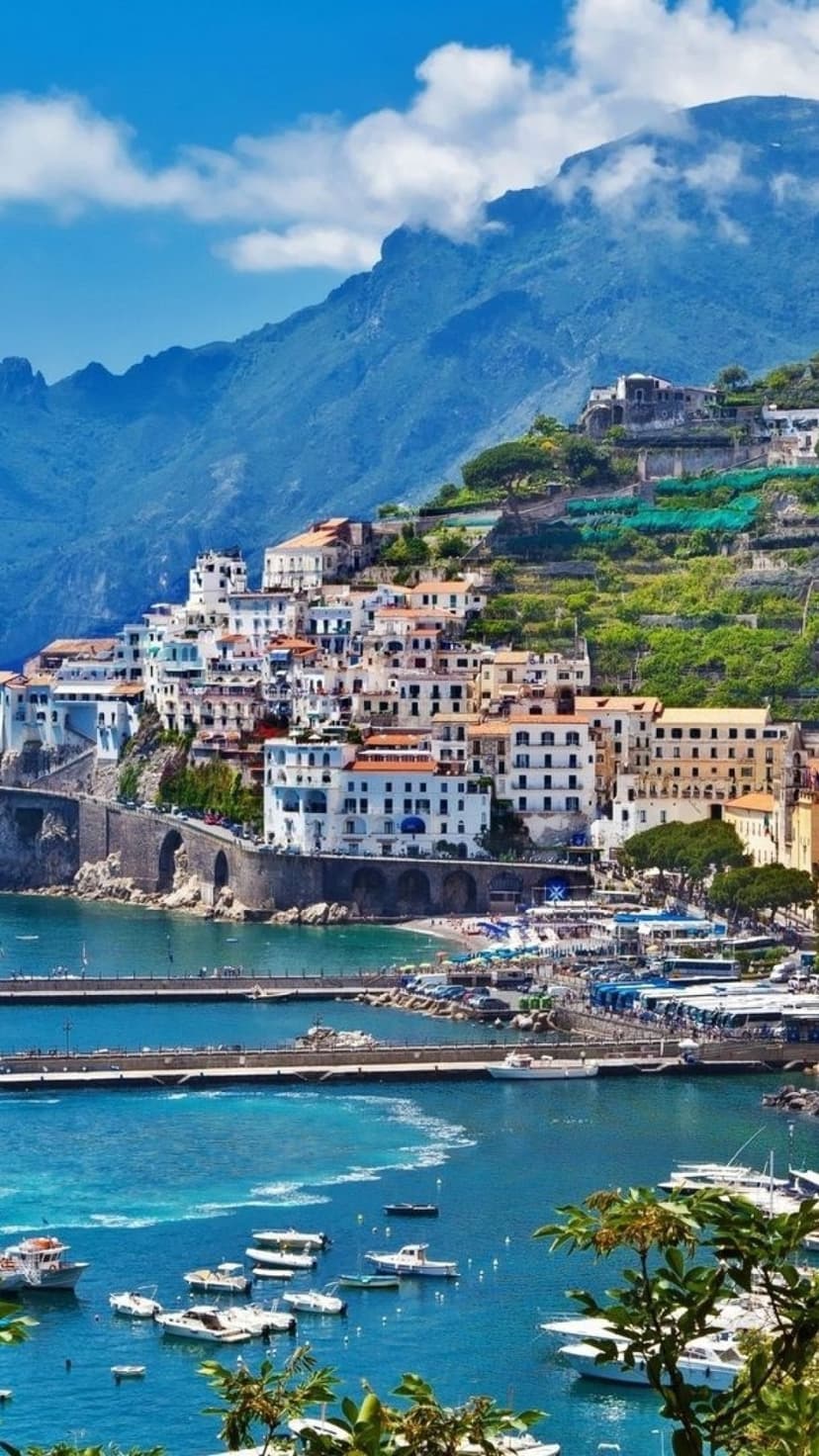 Pastel buildings of an Italian village climb steep cliffs above a turquoise harbor filled with small white boats.