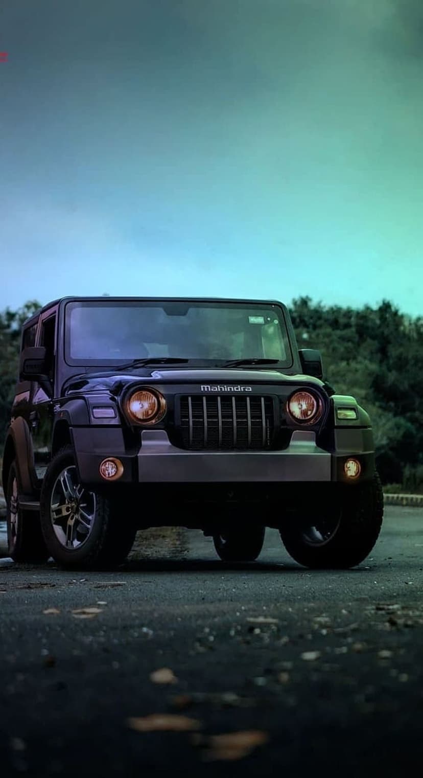 Mahindra Thar SUV in black parked on a paved road with a green and blue sky and dense trees behind it.